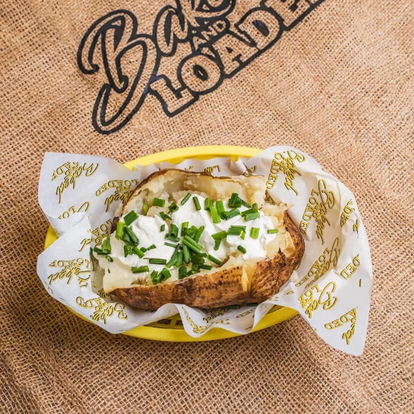 Baked potato with sour cream and chives in a yellow basket on a burlap surface with the restaurant logo.